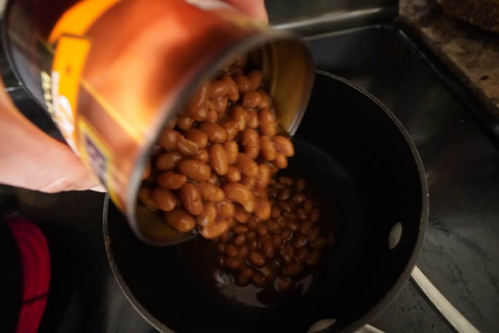 Can of baked beans being poured into a pot on the stove.
