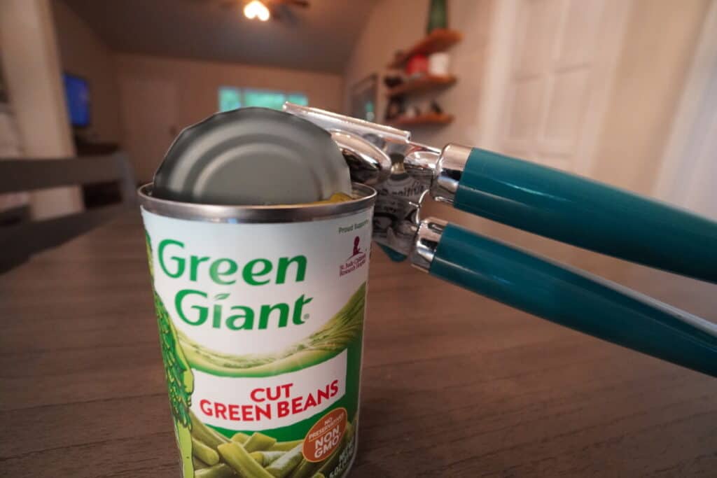 Can of green beans being opened by a can opener on a table.