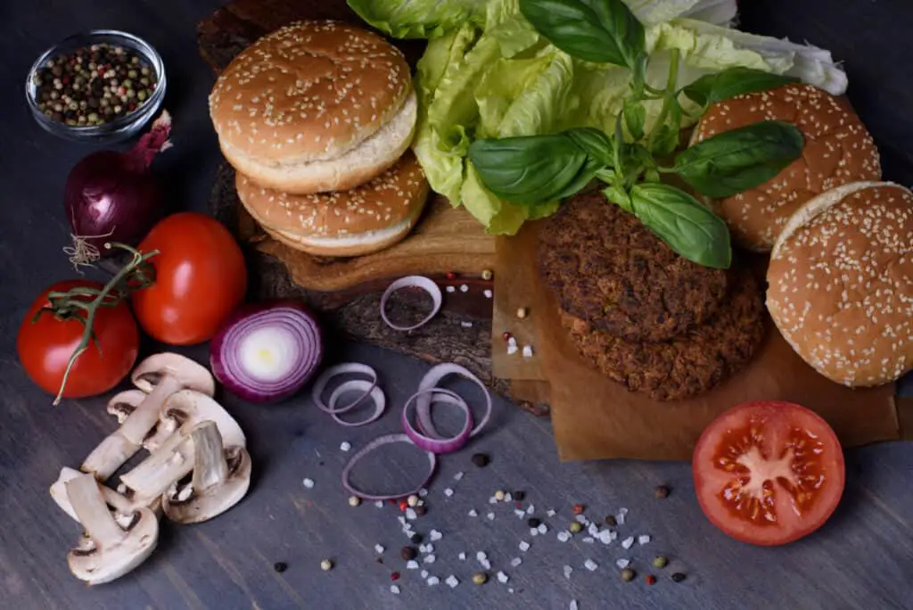 Close up of Burger ingredients: beef patties, sesame bun, fresh vegetables, pepper, mushrooms over dark wooden table.