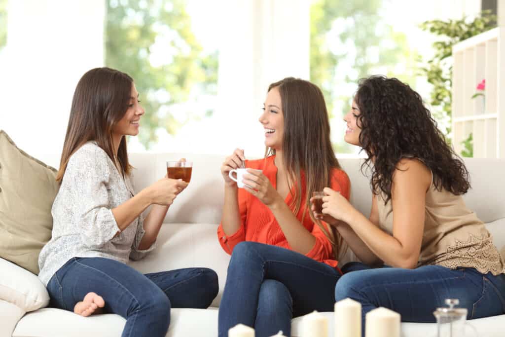 Happy friends talking and drinking coffee and tea sitting on a couch at home