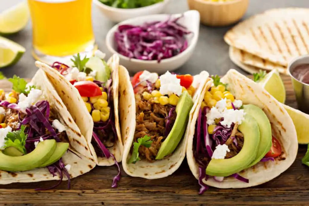 Variety of delicious tacos pulled from a plate next to pork, red cabbage, and avocado