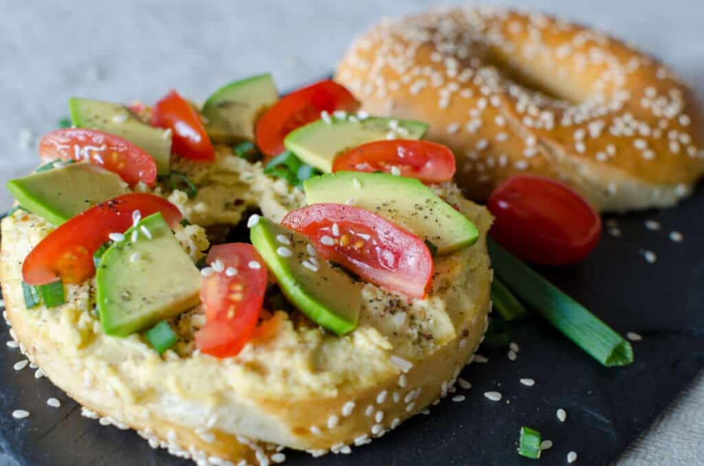 Bagel with hummus, avocado slices, spices, green onion, tomatoes, and sesame seeds