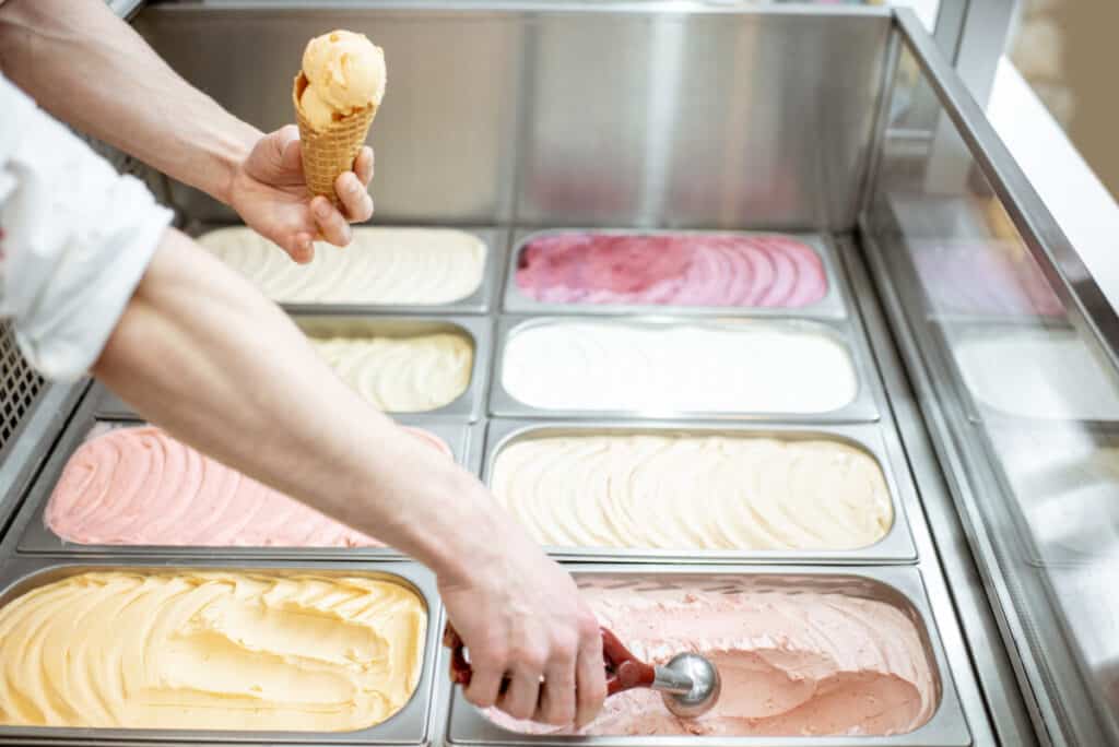 Close up view of male hand with metal trays offering colorful ice cream in waffle cone at display of ice cream refrigerator
