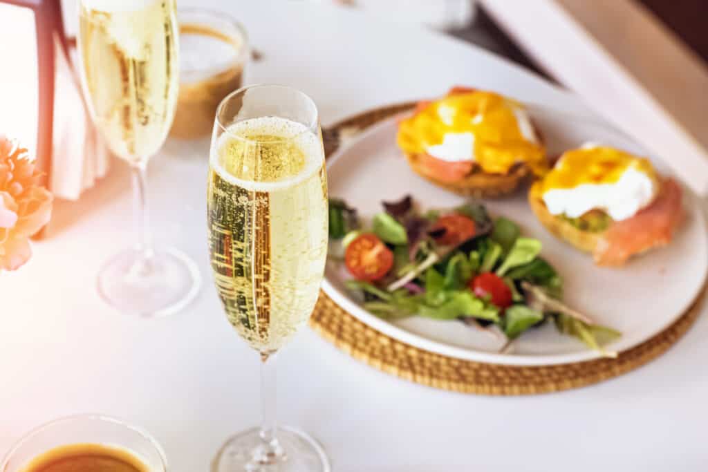 Glasses of champagne and eggs benedict and salad on a white plate on white table