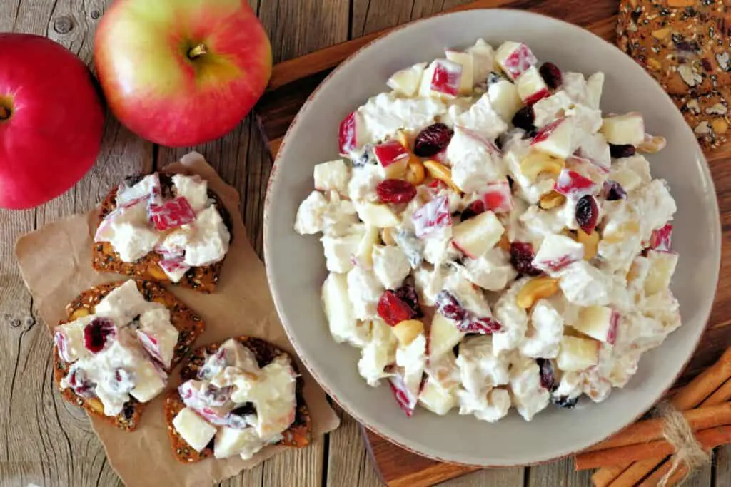 Chicken salad with apples, cranberries, cashews, and crackers in a white bowl on a wooden table.