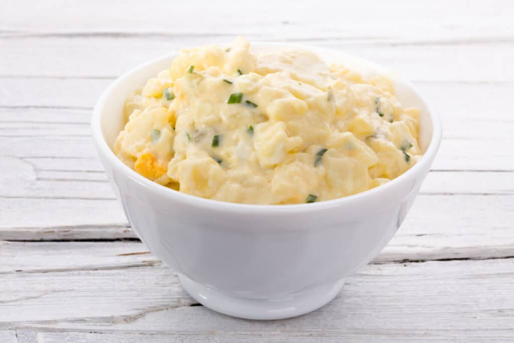 Potato salad in white bowl on white wooden background.